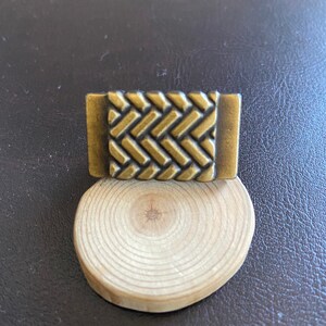 K&ouml;nnte beinhalten: Ein kleines, rechteckiges, messingfarbenes Metallobjekt mit einem gewebten Muster. Das Objekt liegt auf einem Holzsockel.