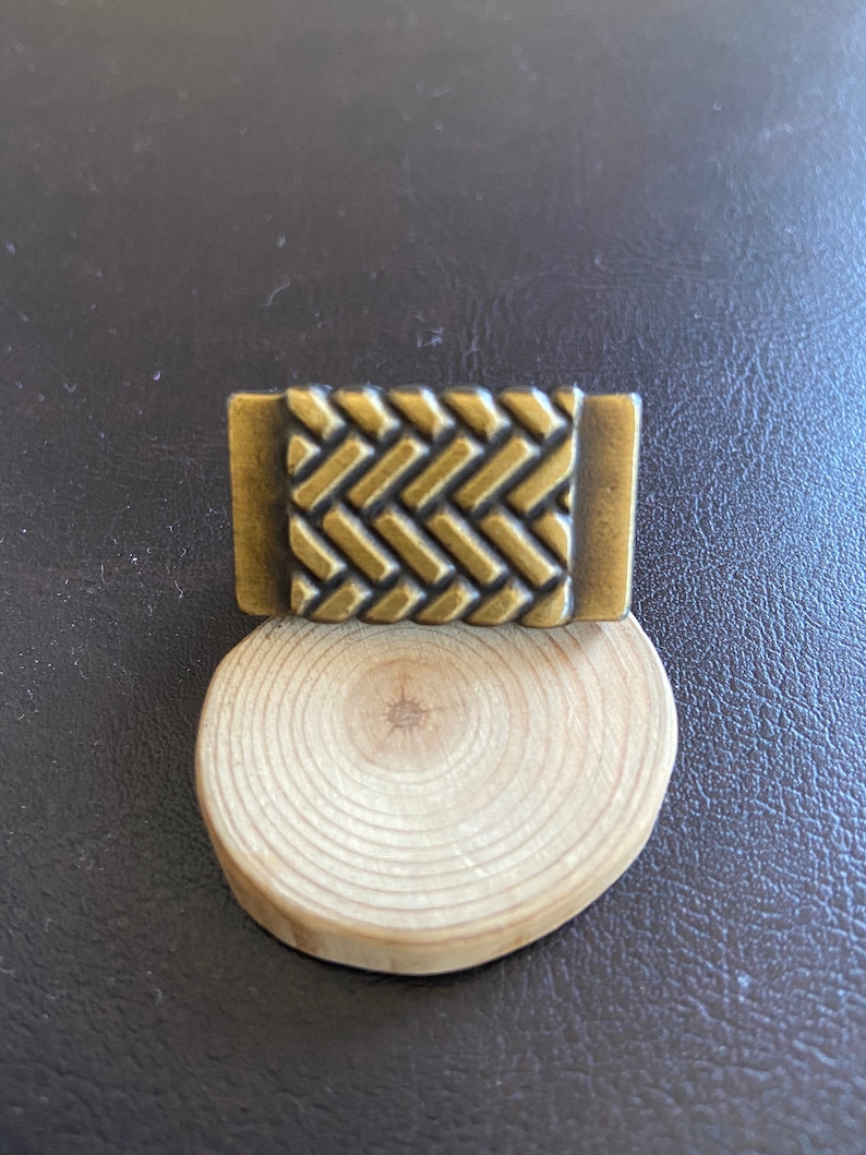 K&ouml;nnte beinhalten: Ein kleines, rechteckiges, messingfarbenes Metallobjekt mit einem gewebten Muster. Das Objekt liegt auf einem Holzsockel.