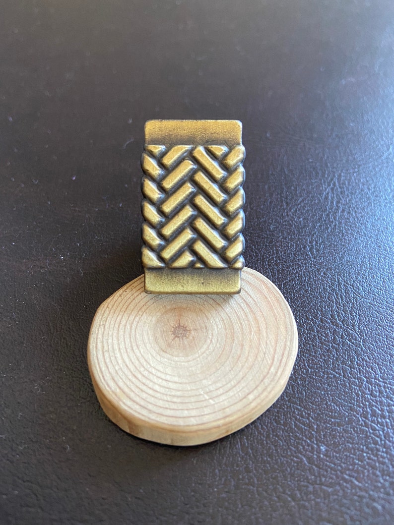 K&ouml;nnte beinhalten: Ein goldfarbenes, dekoratives Metallst&uuml;ck mit einem gewebten Muster. Das St&uuml;ck liegt auf einem Holzsockel.