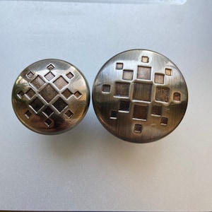 May include: Two antique bronze cabinet knobs with geometric square and diamond patterns. The knobs have a brushed finish and are circular in shape. The design is a mix of recessed and raised elements, creating a textured appearance.