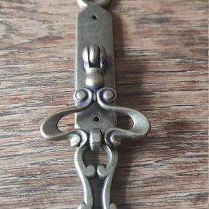 May include: Ornate brass cabinet pull with a decorative design. The pull has a loop handle and a decorative backplate.