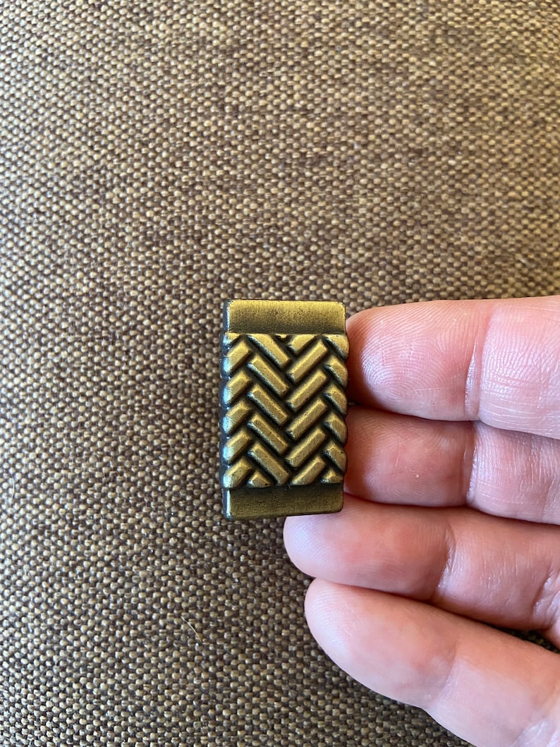 K&ouml;nnte beinhalten: Ein kleiner, rechteckiger, messingfarbener Schubladengriff mit einem gewebten Muster.