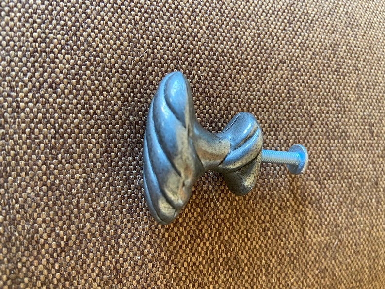 May include: A silver-colored cabinet knob with a decorative, twisted design. The knob is attached to a screw with a blue head. The hardware is likely for a drawer or cabinet.