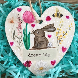 May include: A heart-shaped wooden plaque featuring a painted brown bunny, flowers, and a bee. The plaque has the words "dream big" and is decorated with pink hearts. A jute string is attached for hanging.