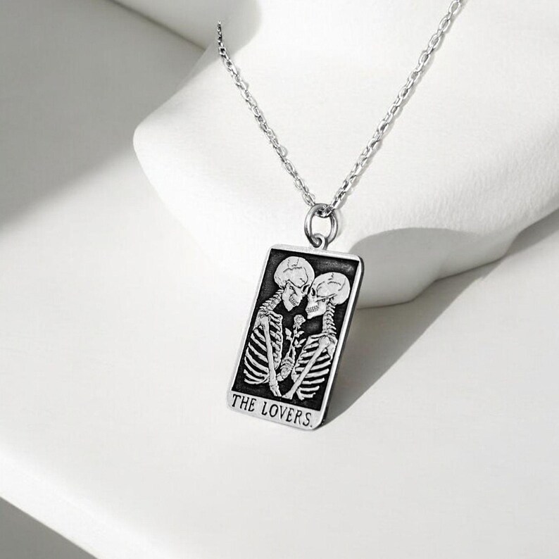 May include: A silver chain necklace with a rectangular pendant featuring a black and white illustration of two skeletons embracing. The pendant has the text "THE LOVERS" inscribed at the bottom.