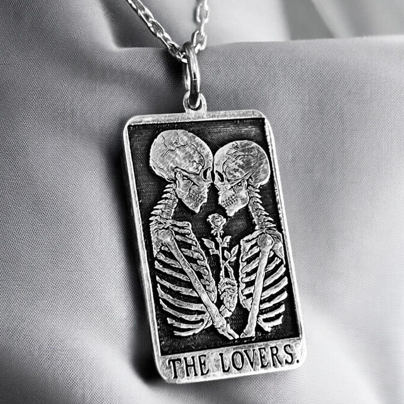 May include: A silver pendant necklace featuring a black and white image of two skeletons embracing with a rose between them. The pendant is rectangular and has the words "THE LOVERS." inscribed at the bottom.