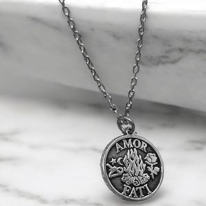 May include: Silver necklace with a round pendant featuring the words "AMOR FATI" and a design of a campfire with a crescent moon, a rose, and a star.