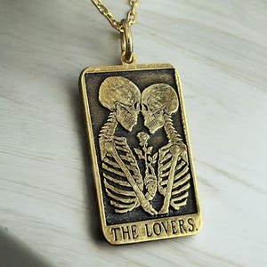 May include: Gold pendant necklace featuring a tarot card design with two skeletons embracing. The card reads "THE LOVERS."