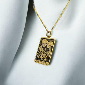 May include: Gold necklace with a rectangular pendant featuring a black and white image of two skeletons embracing. The pendant has the text "THE LOVERS" inscribed at the bottom.