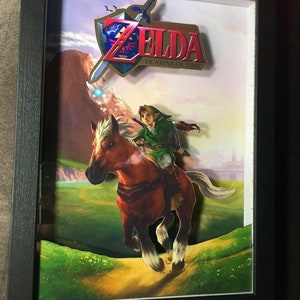 Könnte beinhalten: Ein gerahmtes 3D-Papiermodell von Link, der auf Epona reitet, aus The Legend of Zelda: Ocarina of Time. Das Bild zeigt einen grünen Hintergrund mit einer Bergkette in der Ferne. Der Titel des Spiels ist in goldener und roter Schrift dargestellt.