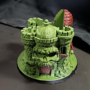 Könnte beinhalten: 3D-gedrucktes Modell eines grünen Schlosses mit schädelartiger Fassade. Das Schloss verfügt über mehrere Türme, einen Turm mit rotem Dach und eine kreisförmige Basis. Das Modell ist detailliert und strukturiert, mit dunklem Hintergrund.