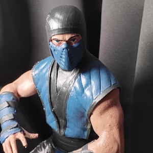 Op de afbeelding: Gedetailleerd beeldje van een personage uit de Mortal Kombat-videogameserie. Het beeldje draagt een blauw en zwart kostuum, inclusief een masker, en heeft een vastberaden uitdrukking. Het beeldje staat in een vechthouding.