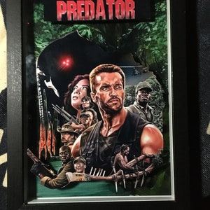 Puede incluir: Una pieza de arte pop-up 3D enmarcada que presenta el póster de la película Predator. El póster presenta a Arnold Schwarzenegger y otros personajes de la película, con el Predator acechando en el fondo. El título "PREDATOR" se muestra en letras rojas en la parte superior del póster.