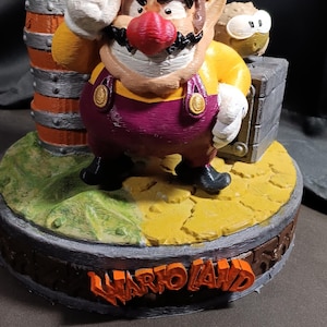 Può includere: Una statuetta di Wario stampata in 3D su una base circolare con la scritta WARIO LAND in arancione. Il personaggio indossa un cappello giallo con una W blu, una maglia gialla, una salopette viola e guanti bianchi. Dietro ci sono un barile e una teiera.