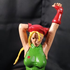 Könnte beinhalten: Handbemalte Figur einer weiblichen Figur in einem grünen Trikot, roten Handschuhen und einem roten Barett. Die Figur hat blondes Haar und posiert mit erhobenen Armen. Ein kleines rotes Herz befindet sich auf der Brust.