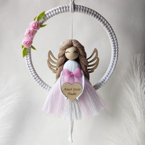 Peut inclure: Un cercle en macramé blanc avec une poupée ange rose et blanche suspendue. L'ange tient un cœur rose avec le texte "Aniol Stróż Nadji" écrit dessus. Le cercle est décoré de fleurs roses.