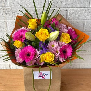 May include: A bouquet of yellow roses, pink gerbera daisies, and white lilies wrapped in orange paper and placed in a brown paper bag with a green handle.