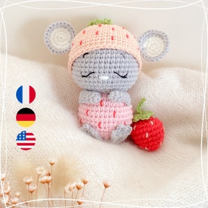 May include: A handmade crochet mouse doll in a strawberry outfit. The doll has grey fur, a pink strawberry hat, and pink strawberry shorts. A red crochet strawberry is placed next to the doll.