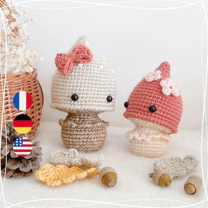 May include: Two handmade crochet mushroom figures. One is off-white with a pink bow, the other is pink with flower accents. Acorns and crochet leaves are scattered around the mushrooms. The mushrooms have black button eyes.