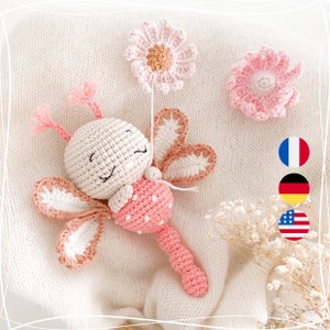 Op de afbeelding: Een gehaakt roze en wit lieveheersbeestje rammelaar speelgoed met een glimlachend gezicht. Het speelgoed heeft een lange handvat en is versierd met twee roze bloemen.