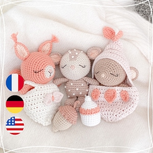 May include: Three crocheted stuffed animals, a deer, a squirrel, and a bear, are lying on a white blanket. The deer is wearing a brown and white polka dot dress, the squirrel is wearing a white dress, and the bear is wearing a pink hooded blanket. The bear is holding a pink bottle.