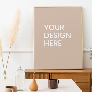 Puede incluir: Un lienzo de color marrón claro enmarcado con el texto "YOUR DESIGN HERE" en blanco. El lienzo está colgado en una pared sobre un armario de madera. Hay un jarrón con hierba de la pampa seca, una botella de vidrio, una pera y una taza de cerámica marrón sobre una mesa blanca delante del armario.