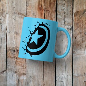 May include: A light blue ceramic mug with a black and white graphic of a superhero shield with a star in the center. The shield is cracked and broken, as if it has been shattered.
