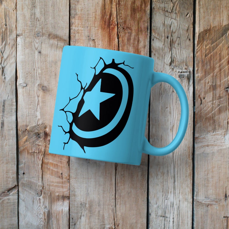May include: A light blue ceramic mug with a black and white graphic of a superhero shield with a star in the center. The shield is cracked and broken, as if it has been shattered.