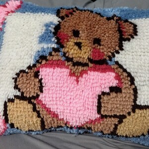 May include: A plush pillow with a brown teddy bear wearing a pink heart-shaped shirt. The pillow is made of a soft, fuzzy material and has a white background with blue accents.