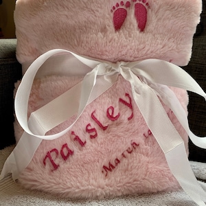 Puede incluir: Una manta rosa y afelpada para bebé con huellas de pies rosas bordadas, el nombre "Paisley" y la fecha "19 de marzo" en rosa. La manta está atada con una cinta blanca.