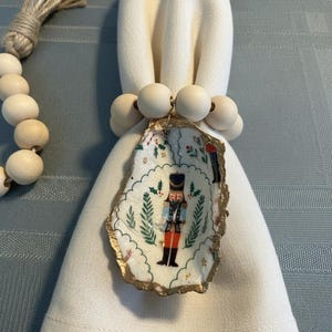 May include: A white linen napkin is adorned with a decorative oyster shell napkin ring. The shell features a painted nutcracker design with gold trim. Wooden beads encircle the napkin. A strand of wooden beads is visible.