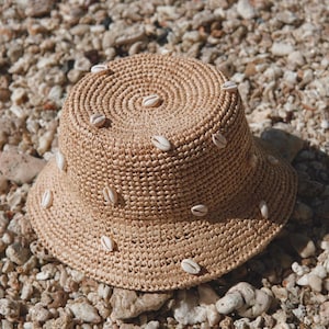 May include: A woven straw bucket hat with a wide brim, adorned with small white seashells. The hat is a light tan color and sits on a bed of pebbles and small rocks. The hat's design is circular, with the seashells evenly spaced.