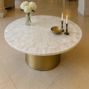 Puede incluir: Una mesa de centro redonda con una superficie blanca en mosaico y una base dorada. Está adornada con un jarrón con flores blancas, un cuenco dorado y dos candelabros negros con velas blancas. La mesa está en una habitación espaciosa y luminosa. El diámetro es de unos 120 cm.