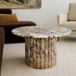 Puede incluir: Una mesa de centro redonda de mármol con una base cilíndrica acanalada. La encimera presenta un patrón arremolinado de blanco y morado. La mesa es un punto focal en una sala de estar contemporánea. El diámetro de la mesa es de unos 90 cm.
