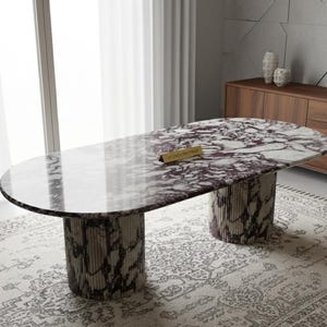May include: Oval marble dining table with a white and purple patterned top. The table is supported by two cylindrical bases with a matching marble design. A small gold sign is on the table. The table is approximately 72 inches long.