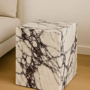 May include: A square marble side table with a white and dark purple veined pattern. The cube-shaped table is made of stone and sits on a wooden floor. The table is next to a beige sofa.