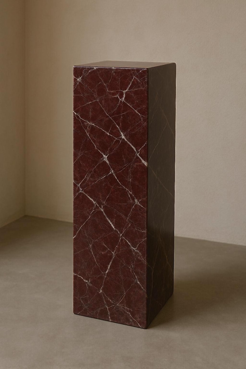 Op de afbeelding: Een hoge, rechthoekige sokkel met een bordeauxrode marmerlook en witte adering. De solide, geometrische vorm is geschikt voor het presenteren van decoratieve items. Het oppervlak heeft een gepolijste uitstraling. Het object staat in een neutrale ruimte.