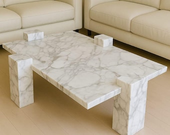 May include: A rectangular marble coffee table with a white and gray veined pattern. The table has four square supports and four rectangular legs. The table is in a modern living room setting.