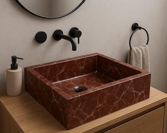May include: A square, red marble bathroom sink with white veining sits atop a wooden vanity. A black faucet, soap dispenser, and towel ring are mounted on the wall. A round mirror is above the sink.