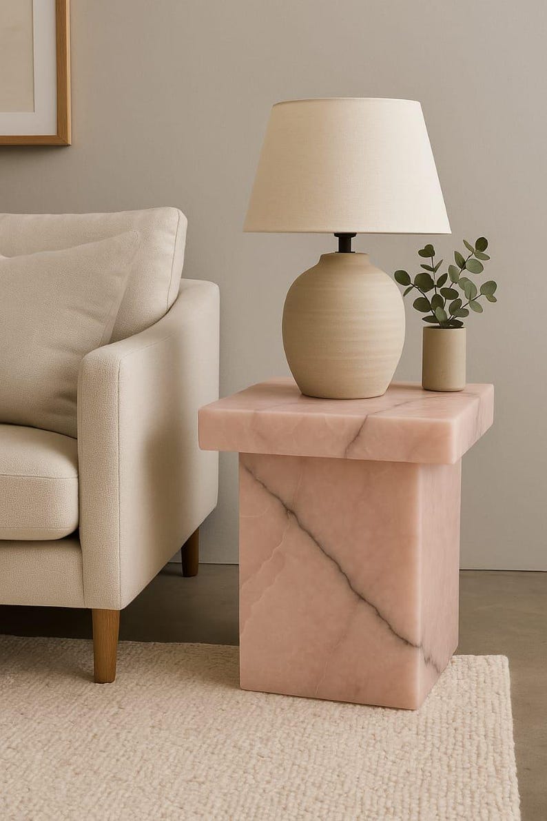 May include: A pink marble side table with a square top and base. A beige lamp with a fabric shade sits on top, next to a small potted plant. The table is next to a beige sofa.