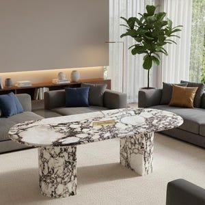 May include: An oval marble coffee table with a white and dark brown pattern. The table rests on two cylindrical bases. The room features gray sofas, blue and brown throw pillows, and a large potted plant.