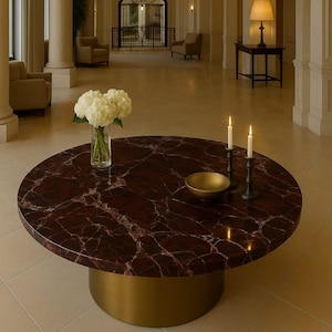 Puede incluir: Una mesa de centro redonda con una tapa de mármol rojo oscuro y una base dorada. La mesa está decorada con un jarrón de flores blancas, un cuenco dorado y dos velas encendidas en candelabros negros. La mesa está en una habitación bien iluminada.