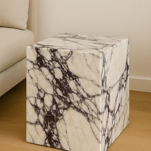 May include: A square marble side table with a white and dark purple veined pattern. The cube-shaped table is made of stone and sits on a wooden floor. The table is next to a beige sofa.