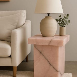 K&ouml;nnte beinhalten: Ein rosa Marmor-Beistelltisch mit quadratischer Ober- und Unterseite. Auf der Oberseite steht eine beige Lampe mit Stoffschirm, neben einer kleinen Topfpflanze. Der Tisch steht neben einem cremefarbenen Sofa.