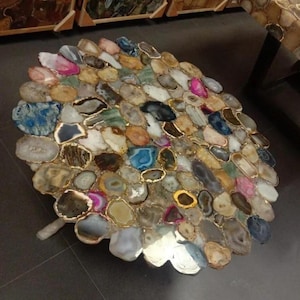 Może przedstawiać: Okrągły blat stołu z agatu o unikalnym wzorze, składający się z wielu różnokolorowych plastrów agatu. Blat stołu wykonany jest z naturalnego agatu i ma polerowane wykończenie.