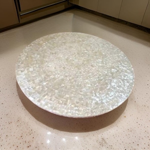 Puede incluir: Una mesa redonda con una superficie de mosaico. La superficie está hecha de pequeñas baldosas rectangulares iridiscentes, creando un efecto brillante. La mesa está sobre un fondo neutro, resaltando su diseño decorativo. La mesa es una pieza decorativa.