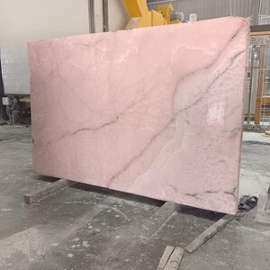 K&ouml;nnte beinhalten: Gro&szlig;e, rechteckige Platte aus rosa Onyx mit dunkelgrauer Maserung. Der Stein wird von zwei grauen Holzbalken getragen. Der Onyx befindet sich in einer Fabrikumgebung mit Industrieanlagen im Hintergrund.