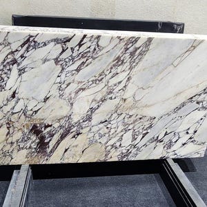May include: A large rectangular marble slab with a white base and intricate veining in shades of purple and black. The stone is displayed on a metal stand, showcasing its polished surface and natural patterns. The slab's dimensions are not visible.