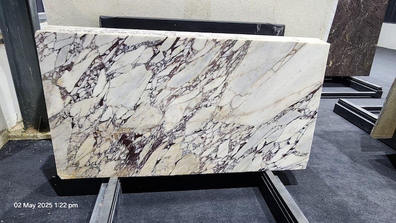 May include: A large rectangular marble slab with a white base and intricate veining in shades of purple and black. The stone is displayed on a metal stand, showcasing its polished surface and natural patterns. The slab's dimensions are not visible.