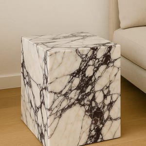 May include: A square marble side table with a white base and dark purple veining. The cube-shaped table is placed on a light wood floor, with a beige sofa in the background. The table is a modern piece of furniture.